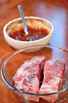 Two pieces of raw pork necks and bowl with spicy marinade on a desk.