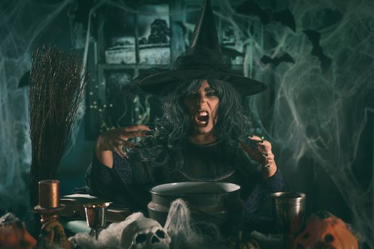 Witch with awfully face and hat on her head in creepy surroundings full of cobweb sends evil.