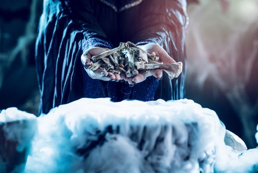 Close-up of a witch's hands holding bones above boiling water for a cooking poison soup.