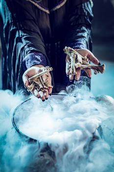 Close-up of a witch's hands holding bones above boiling water for a cooking poison soup.