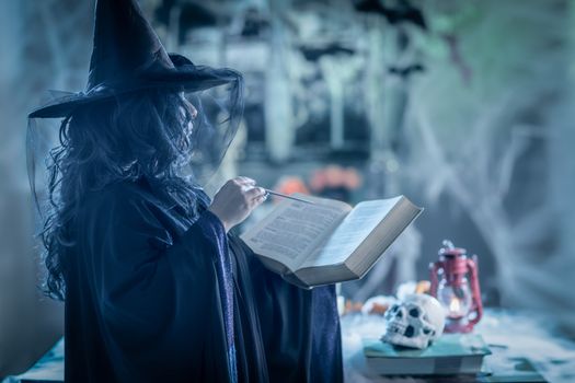 Witch in black cloak with long black hair and black hat in creepy surroundings  reading recipe of magic potion.