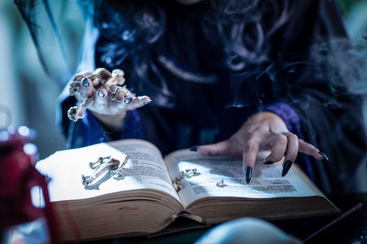 Close up of a witch's hands with pointed black nails holding bones above book of a magic recipe.