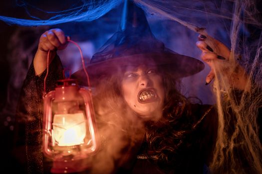 Portrait of witch with awfully face and lighted lantern in her hand in creepy foggy surroundings sends evil.