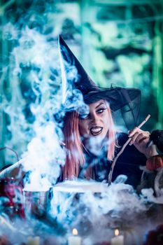 Witch with smiling face in creepy surroundings cooks poison soup in boiling cauldron.