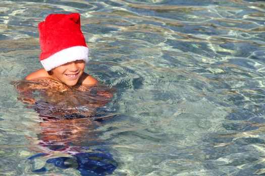 Portrait of cute smiling boy in Santa hat splashing in beautiful transparent tropical sea, christmas sea vacation concept