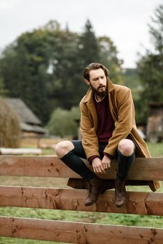 man in a brown coat and torn pants sitting on a wooden fence