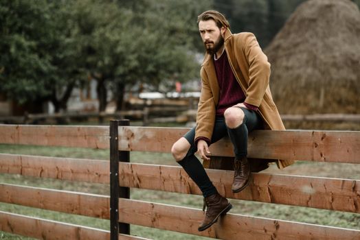 man in a brown coat and torn pants sitting on a wooden fence