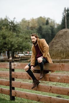 man in a brown coat and torn pants sitting on a wooden fence