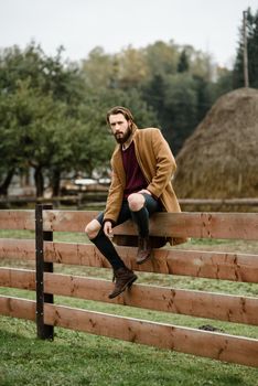 man in a brown coat and torn pants sitting on a wooden fence