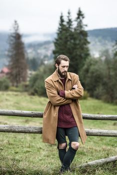 man in a brown coat and torn trousers was leaning against the fence