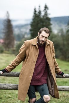 man in a brown coat and torn trousers was leaning against the fence