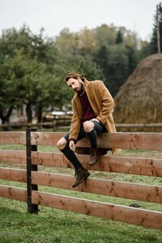 man in a brown coat and torn pants sitting on a wooden fence