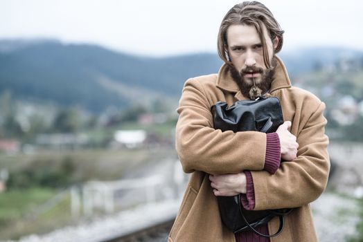 a man in a brown coat stands clutching a black briefcase on a background of mountains