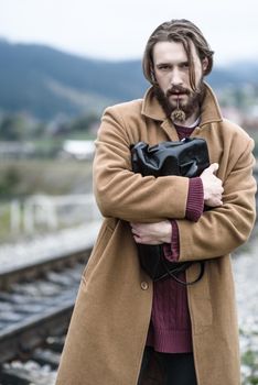 a man in a brown coat stands clutching a black briefcase on a background of mountains