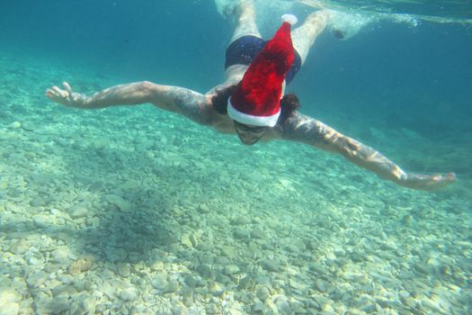 Man in Santa Claus hat swimming underwater in sea, Christmas vacation at sea concept