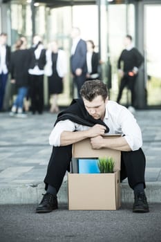 Fired business man sitting frustrated and upset on the street near office building with box of his belongings. He lost work