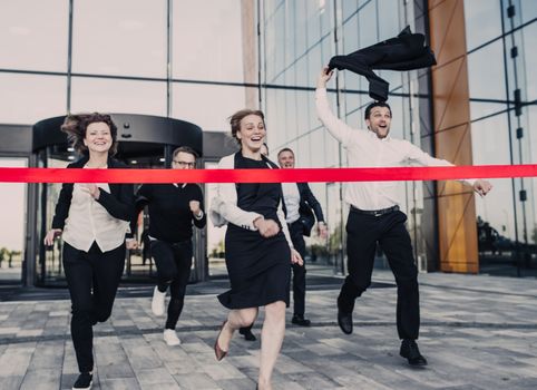 Group of happy business people running from office building crossing red ribbon finish line