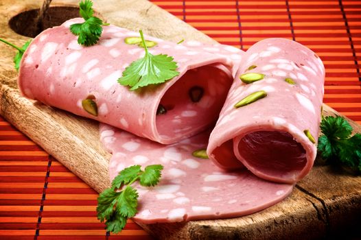 Slices of Delicious Fresh Mortadella with Pistachio and Greens Rolled Up on Wooden Cutting Board closeup on Straw mat background