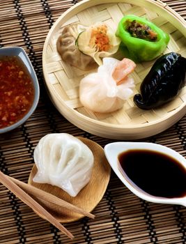 Assorted Dim Sum in Bamboo Steamed Bowl and Ika with Shrimps on Wooden Plate with Red Chili and Soy Sauces and Chopsticks closeup on Straw Mat background