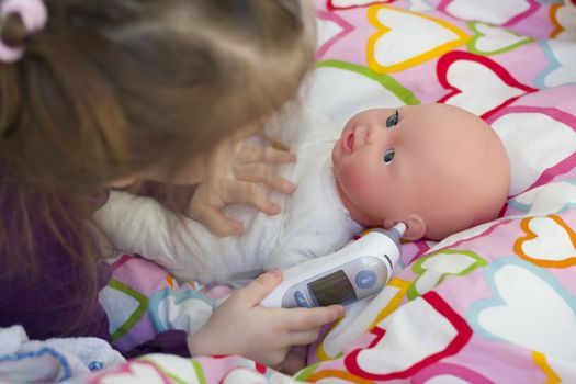 Little girl playing doctor with a doll, measuring temperature with electronic themometer and taking care of a doll, concept maternity, lifestyle and childhood