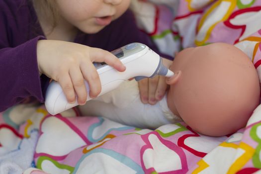 Little girl playing doctor with a doll, measuring temperature with electronic themometer and taking care of a doll, concept maternity, lifestyle and childhood