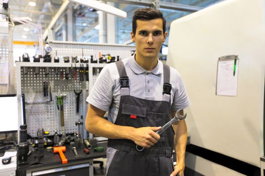 Portrait of a young worker holding wrench at workplace with computer and tools at CNC factory