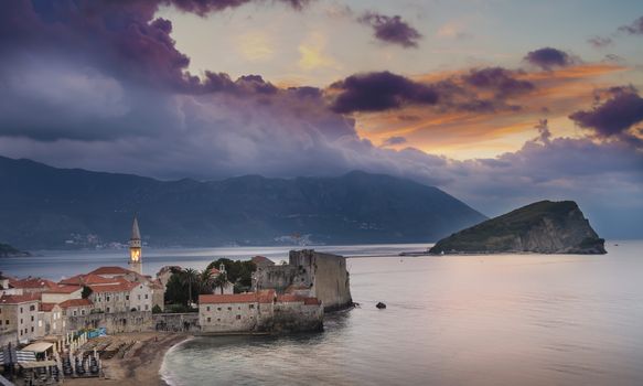 Sunrise over Budva old town with lit tower of Sv. Ivan church, old town's citadel and island Sv. Nikola to the right. Picturesque mountains and coast in background