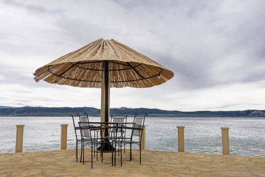 Terrace at the sea with chairs, table and straw parasol, sea and an island in the background