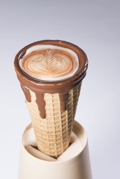 Coffee in waffle cone dipped in chocolate with drawings in capuccino / latte milk foam, latte art, isolated