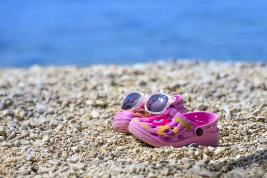 Pink beach kid's crocs and sunglasses on sandy beach.Beach sandals and sunglasses in the foreground and blurred sea in the background.