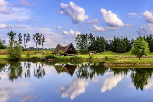 A small traditional cottage covered with straw at a lake, beautiful reflection of clouds in the water, at the far shore people are fishing (face not recognizable)