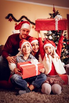 Beautiful happy family wearing santa hat and holding many christmas gifts at the home. Looking at camera.