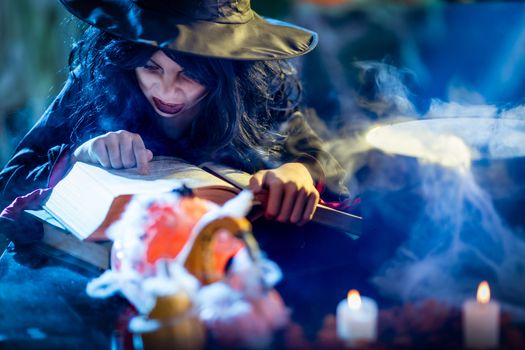 Witch with awfully face reading recipes of magic drink in creepy surroundings and smoky background.