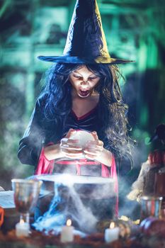 Portrait of young angry witch with awfully face in creepy surrounding watching the future in magic ball above boiling potion. 