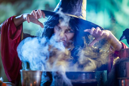 Close-up of young witch with awfully face in creepy surroundings full of steam and fog, tells evil words to magic potion.