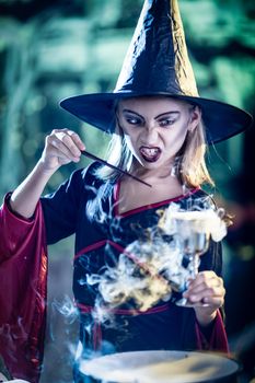 Young witch with angry evil face holds a goblet with magic potion in one and magic wand in the other hand. 