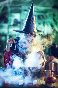 Young witch with face hidden in steam and fog, sends magic to magic potion in cauldron in front her.