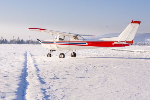 Small sport airplane with white fuselage and red and blue stripes on the apron. The airport is covered in fresh snow on a sunn day.