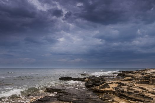 Dramatic skies over a rocky beach and sail boats in distance on horizon