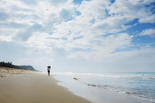 INDIA. VARKALA — JANUARY 27, 2017 : A lonely man with a load on his head leaving the camera along the beach along the ocean with small calm waves and haze on the horizon. Varkala town in the south of India in the state of Kerala, known for its spas.