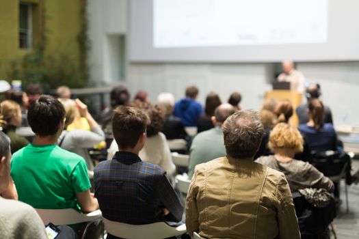 Male speaker giving presentation in lecture hall at university workshop. Audience in conference hall. Rear view of unrecognized participant in audience. Scientific conference event.