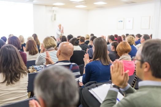 Life coaching symposium. Female speaker giving interactive motivational speech at entrepreneurship workshop. Audience in conference hall. Rear view of unrecognized participant.