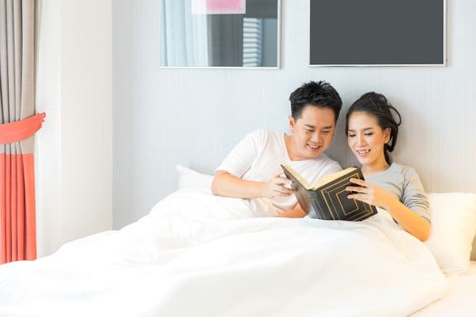 Young Asian Couple laid in a bed reading a book in bedroom of contemporary house for modern lifestyle concept