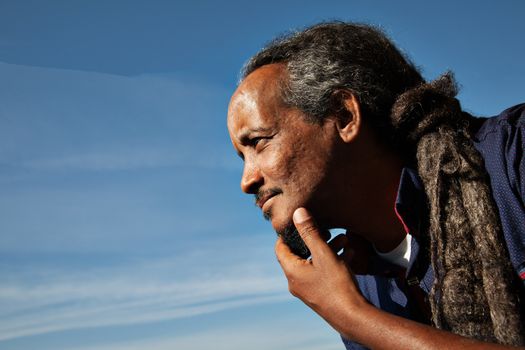 A portrait of a black rastafarian man over a blue sky with some clouds.