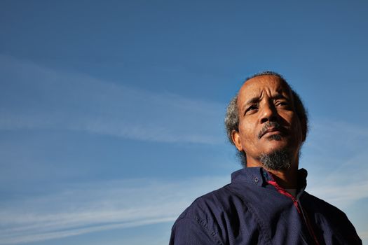 A portrait of a black rastafarian man over a blue sky with some clouds.