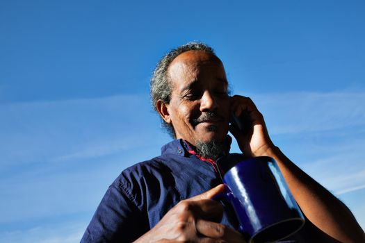 A portrait of a black rastafarian man over a blue sky with some clouds, holding a mug of tea or cofee, on the phone