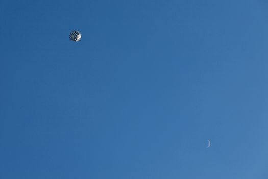 Black and white balloon in flight and moon seen from below against a blue sky