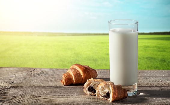 Glass of milk on a rustic table, beside croissants, blurred background with grass and sky