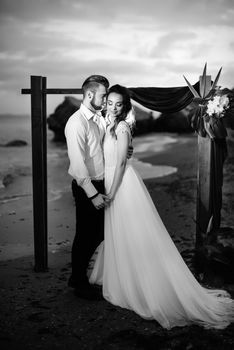 young couple groom and bride with a bouquet in the evening on the beach near the wedding arch