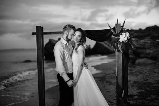 young couple groom and bride with a bouquet in the evening on the beach near the wedding arch